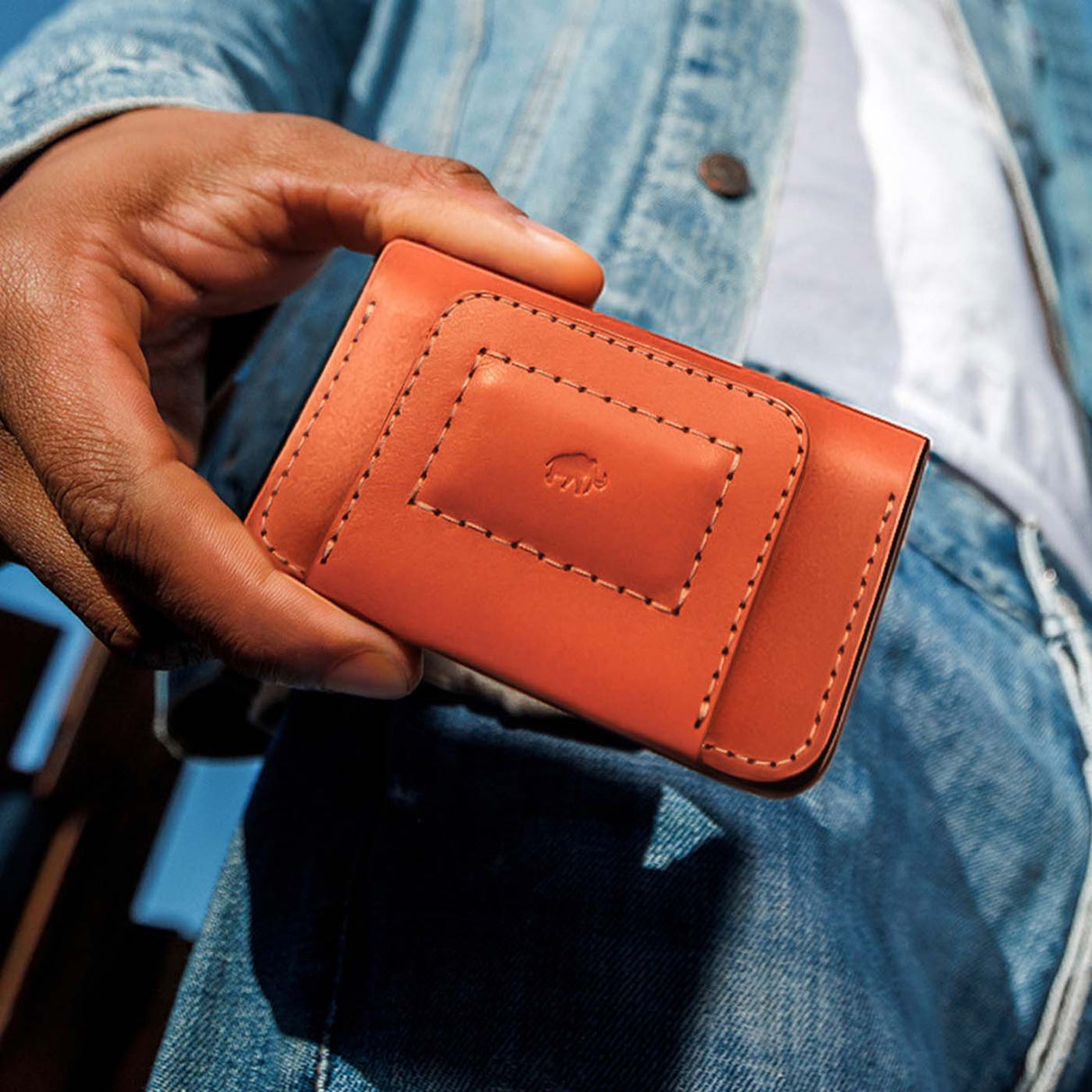 Hand holding a orange leather wallet with a brand logo against a denim background