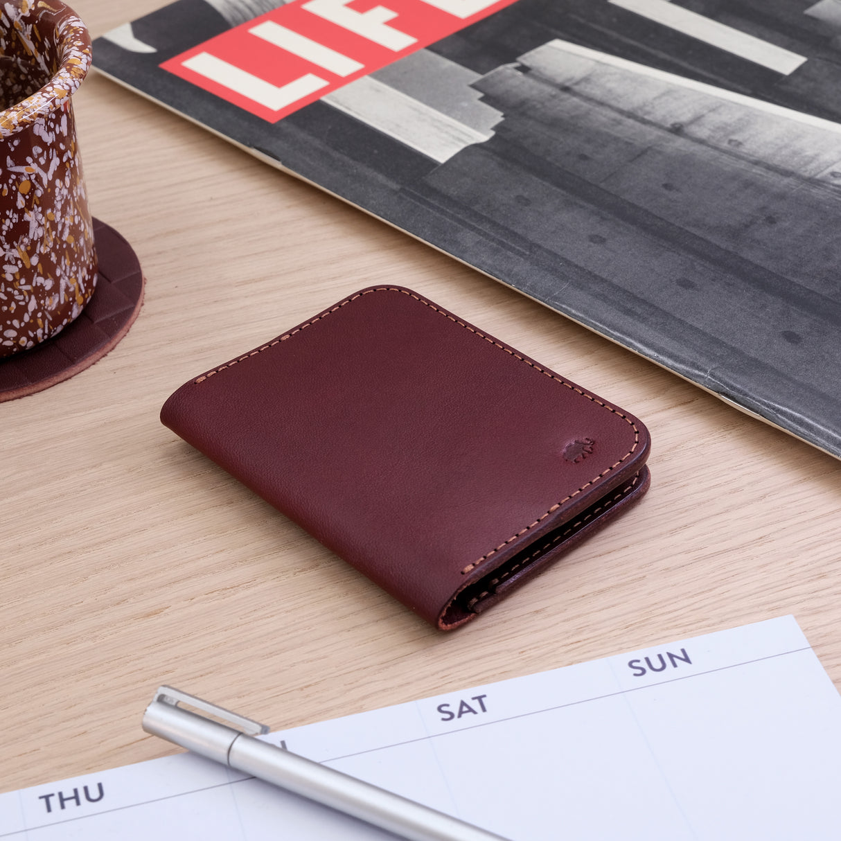 Woolly Made Portrait Wallet in Burgundy. On a wooden table near a coffee mug and magazine.