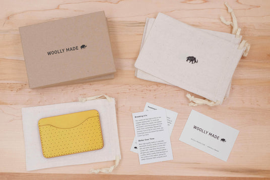 Top down view of Wooly Made packaging including a sustainable brown box, drawstring canvas bags, Woolly Made business and wear and care cards, and a yellow leather Slim Wallet with decorative perforation.