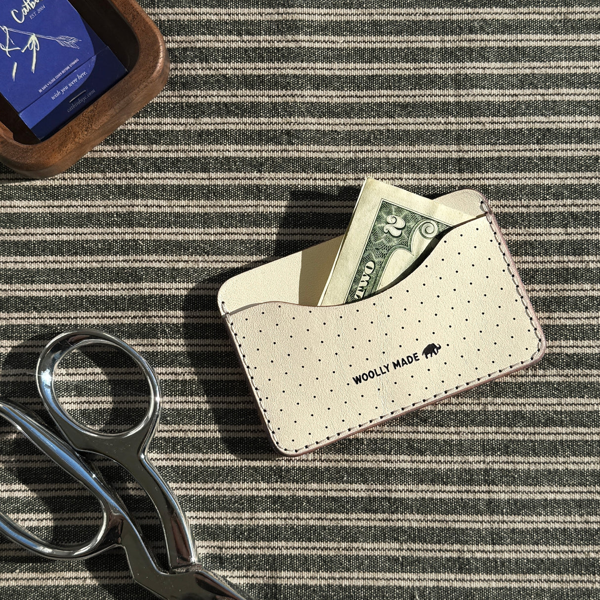 Woolly Made Slim Wallet in Tusk on a green and ivory striped fabric. A $2 bill is coming slightly out of the wallet and a pair of scissors and a wooden tray with Catbird matches are near the wallet.