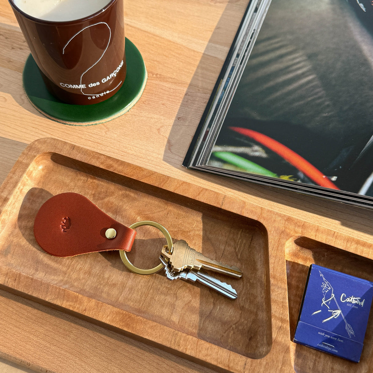 Woolly Made Brown Tab Keychain on a wooden table near a Comme Des Garcons Candle and a open magainze.