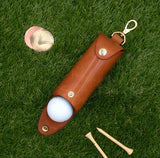 Golf ball in a leather case on grass with a tee and ball marker.