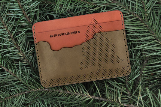 Top down view of the brown and olive leather Keep Forests Green Half Wallet with Oregon state-shaped pocket, perforated fir tree design, and Woolly Made logo on top of fir tree brances.