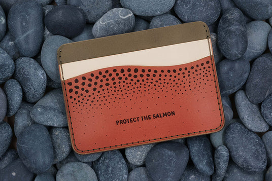 Top down view of the olive, tusk, and salmon leather Protect the Salmon Half Wallet with salmon skin design on top of a bed of rocks.