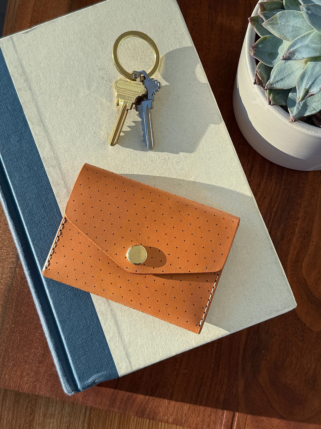 Woolly Made tan Snap Wallet lays ontop of a ivory and blue book with keys nearby. There a cute succulent plant next to the book.